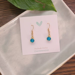 May include: A pair of gold wire wrapped earrings with blue glass beads. The earrings are displayed on a white card with the text "Handmade Jewelry Charmbangles".