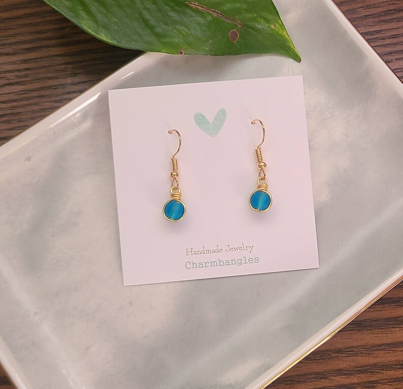 May include: A pair of gold wire wrapped earrings with blue glass beads. The earrings are displayed on a white card with the text "Handmade Jewelry Charmbangles".