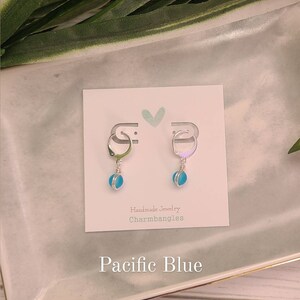 May include: A pair of silver hoop earrings with a light blue glass charm hanging from each hoop. The earrings are displayed on a white card with the text "Handmade Jewelry Charmbangles" and "Pacific Blue".