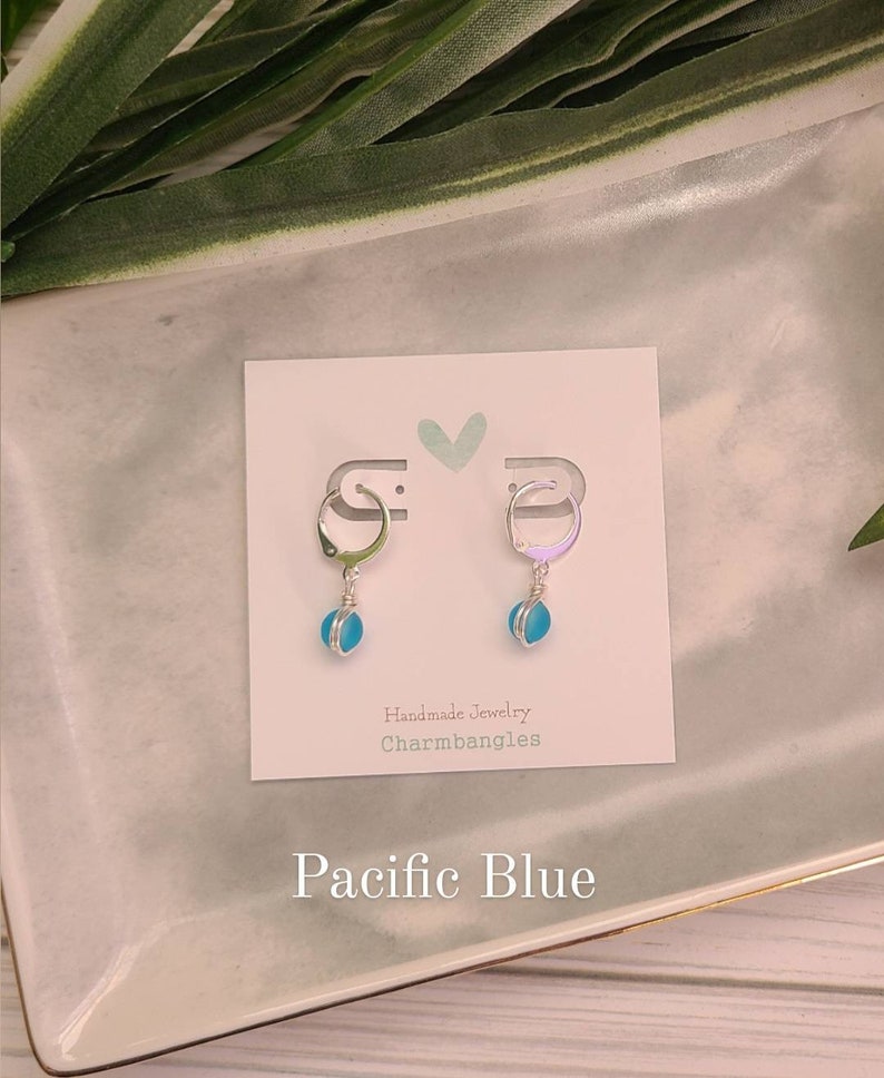May include: A pair of silver hoop earrings with a light blue glass charm hanging from each hoop. The earrings are displayed on a white card with the text "Handmade Jewelry Charmbangles" and "Pacific Blue".