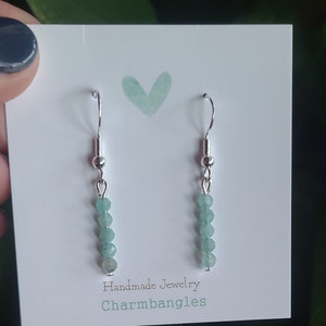 May include: A pair of silver dangle earrings with light green gemstone beads. The earrings are on a white card with the text "Handmade Jewelry Charmbangles".