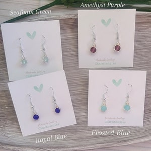 May include: Four pairs of handmade earrings with different colored glass beads. The colors are seafoam green, amethyst purple, royal blue, and frosted blue. Each pair is displayed on a white card with the color name and "Handmade Jewelry Charmbangles" written on it.