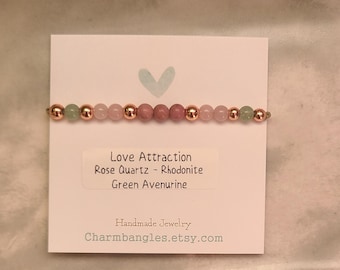 May include: A pink, green, and rose gold beaded bracelet with a card that reads "Love Attraction Rose Quartz - Rhodonite Green Avenurine Handmade Jewelry Charmbangles.etsy.com"