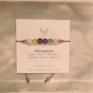 May include: A gray string bracelet with seven gemstone beads: white howlite, yellow citrine, brown smoky quartz, purple amethyst, green fluorite, and white moonstone. The bracelet is labeled "Menopause" with the stones listed below. The card also says "Handmade Jewelry" and "Charmbangles.etsy.com".