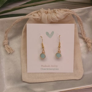 May include: A pair of gold dangle earrings with light blue round glass beads. The earrings are displayed on a white card with the text "Handmade Jewelry Charmbangles".