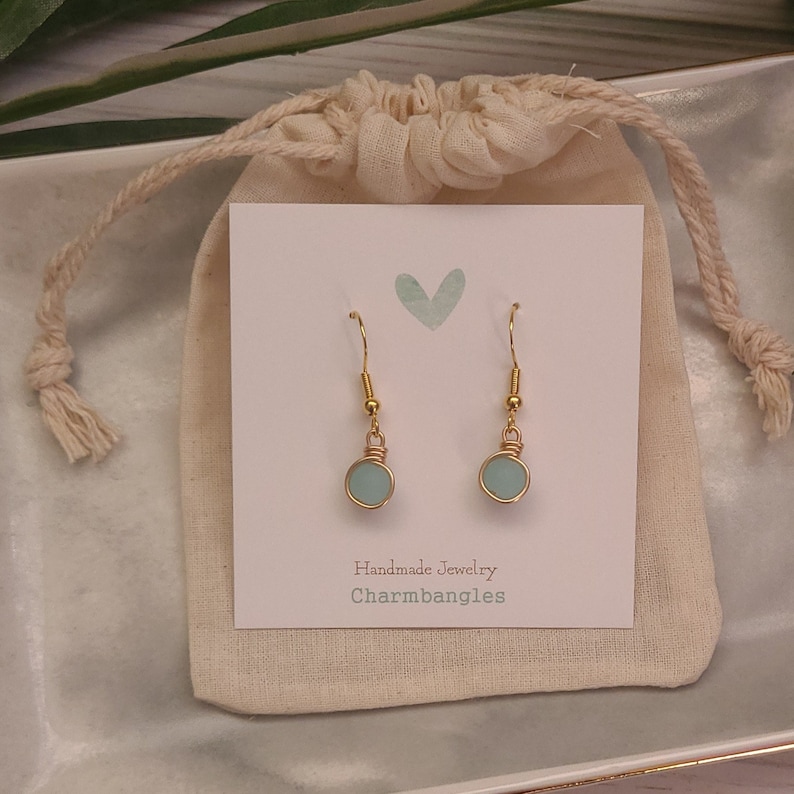 May include: A pair of gold dangle earrings with light blue round glass beads. The earrings are displayed on a white card with the text "Handmade Jewelry Charmbangles".