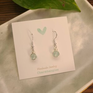 May include: A pair of silver dangle earrings with light blue round gemstone beads. The earrings are displayed on a white card with the text "Handmade Jewelry Charmbangles".