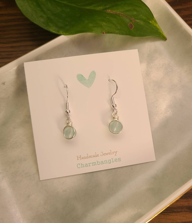 May include: A pair of silver dangle earrings with light blue round gemstone beads. The earrings are displayed on a white card with the text "Handmade Jewelry Charmbangles".