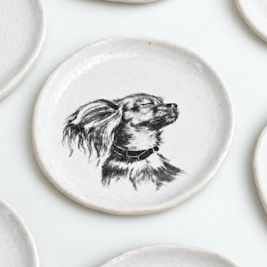 Pode incluir: Uma coleção de pratos redondos de cerâmica em tom branco sujo com texturas salpicadas, cada um apresentando uma ilustração detalhada em preto e branco estilo tinta de diferentes raças de cães, incluindo um pug, um corgi e um chihuahua.