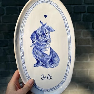 Könnte beinhalten: Eine ovale Keramikplatte mit weißer Glasur und einer handgemalten blauen Illustration eines struppigen Hundes mit Halstuch. Über dem Hund befindet sich ein kleines blaues Herz und darunter der Name Belle in blauer Schreibschrift. Ein blauer Zierrand umrahmt den Rand.