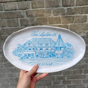 Peut inclure: Un plat ovale en céramique blanche orné d'une illustration peinte à la main en bleu représentant une grande maison avec une tourelle. Le texte "The Lockharts" est écrit en bleu au-dessus et la date "08.08.2026" est inscrite en dessous.