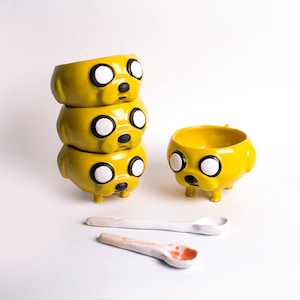 May include: A stack of three yellow ceramic bowls shaped like a cartoon dog with black eyes and a black nose. The bowls are stacked on top of each other, with the largest bowl on the bottom and the smallest bowl on top. There is a fourth bowl in the background, also yellow and shaped like a cartoon dog. The bowls are on a white surface.
