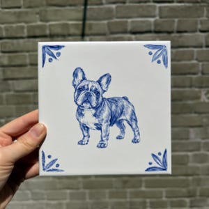 Op de afbeelding: Een vierkante witte keramische tegel met een gedetailleerde blauwe inktillustratie van een Franse Bulldog die in het midden staat. De tegel is versierd met blauwe florale accenten in elk van de vier hoeken, wat zorgt voor een gebalanceerd ontwerp.