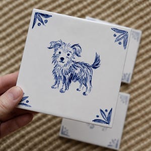 May include: A square white ceramic tile featuring a hand-painted blue illustration of a small, shaggy dog in the centre. The tile has decorative blue floral-inspired corner accents. The design is rendered in a brushstroke style on a smooth surface.