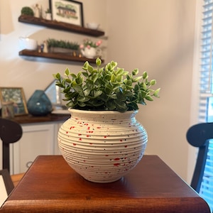May include: A round, white ceramic planter with horizontal ridges and red paint speckles, containing a small, bushy green artificial plant with numerous oval-shaped leaves. The planter has a wide, rounded body and a slightly flared rim.