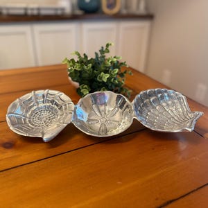 May include: A silver-toned metal serving dish featuring three connected compartments shaped like different seashells. Each section displays distinct embossed textures, including a sand dollar pattern, a scallop shell, and a radial shell design.