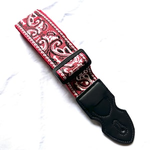 Op de afbeelding: Een gitaarband met een rood en wit paisley-patroon op een geweven stoffen basis. De band is voorzien van een zwarte plastic verstelbare schuif en een zwart lederen uiteinde met versterkte stiksels en een centraal gat voor bevestiging.