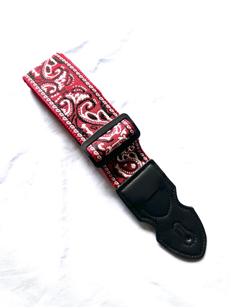 Op de afbeelding: Een gitaarband met een rood en wit paisley-patroon op een geweven stoffen basis. De band is voorzien van een zwarte plastic verstelbare schuif en een zwart lederen uiteinde met versterkte stiksels en een centraal gat voor bevestiging.