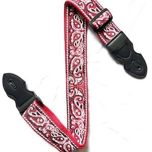 Op de afbeelding: Een gitaarband met een rood, zwart en wit paisley-patroon. De band is voorzien van zwarte lederen uiteinden met verstevigde stiksels en een verstelbare gesp van zwart kunststof, ontworpen om een muziekinstrument stevig vast te houden.