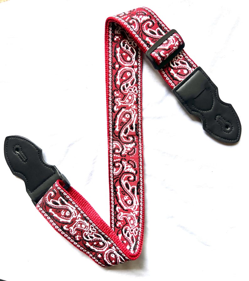 Op de afbeelding: Een gitaarband met een rood, zwart en wit paisley-patroon. De band is voorzien van zwarte lederen uiteinden met verstevigde stiksels en een verstelbare gesp van zwart kunststof, ontworpen om een muziekinstrument stevig vast te houden.