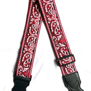 Op de afbeelding: Een gitaarband met een levendig rood, wit en zwart paisley-patroon geweven in de stof. De band bevat een verstelbare zwarte kunststof schuifgesp en twee zwarte lederen uiteinden met verstevigde stiksels en sleufgaten voor bevestiging.