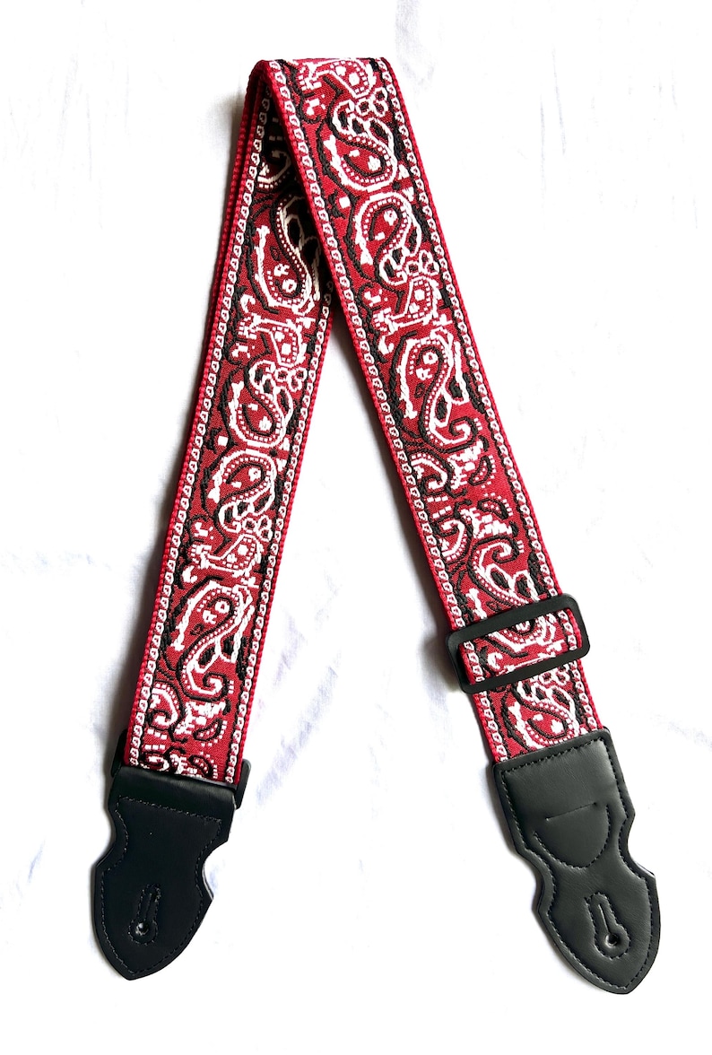 Op de afbeelding: Een gitaarband met een levendig rood, wit en zwart paisley-patroon geweven in de stof. De band bevat een verstelbare zwarte kunststof schuifgesp en twee zwarte lederen uiteinden met verstevigde stiksels en sleufgaten voor bevestiging.