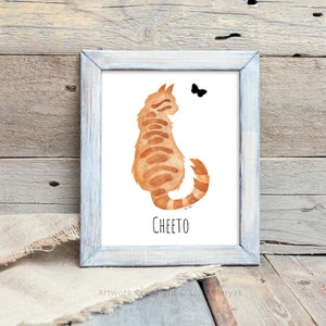 May include: A framed watercolor painting of a ginger cat sitting with its back to the viewer. The cat is looking at a small black butterfly. The painting is titled "Cheeto".