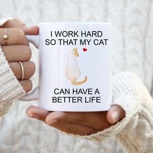 May include: White ceramic mug with a black and white illustration of a cat and the text "I work hard so that my cat can have a better life".