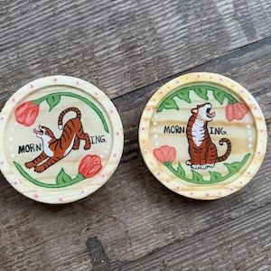May include: Two round wooden coasters with a hand-painted tiger design. Each coaster features a tiger, red flowers, and the word "MORNING." The coasters have a light yellow border with pink dots and green leaves.