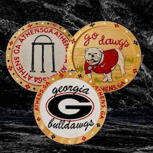 May include: Three wooden coasters with a Georgia Bulldogs theme. One coaster features the Georgia 'G' logo, another has the phrase 'go dawgs' with a bulldog illustration, and the third displays the Athens, GA arch. Each coaster has a decorative border.