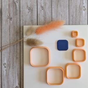 Peut inclure: Un ensemble de six emporte-pièces carrés de différentes tailles, allant du petit au grand. Les emporte-pièces sont en plastique de couleur pêche clair et sont disposés sur une surface blanche. Un petit carré bleu foncé est également inclus dans l'ensemble.