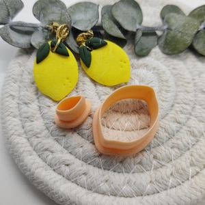 Peut inclure: Boucles d'oreilles jaunes en forme de citron avec des feuilles vertes et des crochets dorés. Deux découpoirs en pâte polymère orange sont également présents. Les boucles d'oreilles et les découpoirs sont sur une surface tissée blanche, avec des feuilles vertes en arrière-plan.
