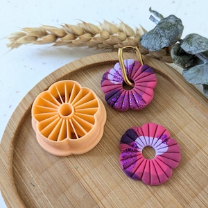 Peut inclure: Une boucle d'oreille en argile en forme de fleur et une fleur en argile assortie avec un crochet de boucle d'oreille doré. Les boucles d'oreilles sont roses, violettes et blanches. Un emporte-pièce en argile orange en forme de fleur est également présent. Les articles sont sur un plateau en bois.