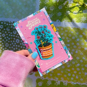 May include: A pink and blue sticker with a potted plant and the text "May the flowers remind us why it was necessary to sit in the rain". The sticker is on a pink background with the text "Harvest Moon Gemz" at the top.