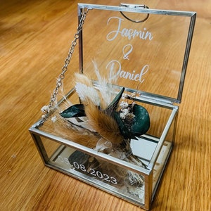 Könnte beinhalten: Eine Glasbox mit einer silbernen Kette und einem durchsichtigen Deckel. Die Box ist mit getrockneten Blumen und Grün gefüllt. Der Deckel trägt die Namen "Jasmin & Daniel" und das Datum "05.08.2023".