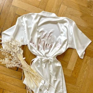 May include: A white satin bridal robe and romper set. The robe features the words "Bride Nur" in a copper-colored script with floral accents. The set is tied with a matching satin belt. A bundle of dried flowers rests beside the set.