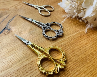 May include: Three pairs of decorative scissors with ornate handles. The scissors are gold, silver, and black. The scissors are on a wooden surface.