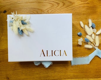 May include: A white gift box with a blue and white floral arrangement on the top. The box is decorated with the name "Alicia" in gold lettering. A blue ribbon is tied around the box.