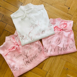 May include: Three satin robes in white and pink, tied with bows. The white robe has "Braut Emma" printed on it, while the pink robes say "Team Braut Antonia" and "Team Braut Mieke". Floral accents surround the text.
