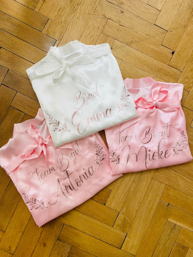 May include: Three satin robes in white and pink, tied with bows. The white robe has "Braut Emma" printed on it, while the pink robes say "Team Braut Antonia" and "Team Braut Mieke". Floral accents surround the text.