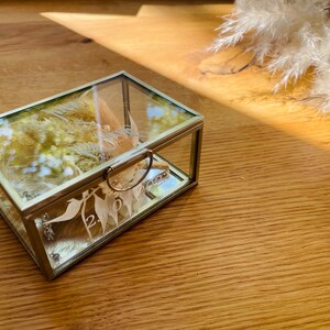 May include: A glass ring box with a gold trim and a floral design. The box is open and the inside is lined with white fabric.