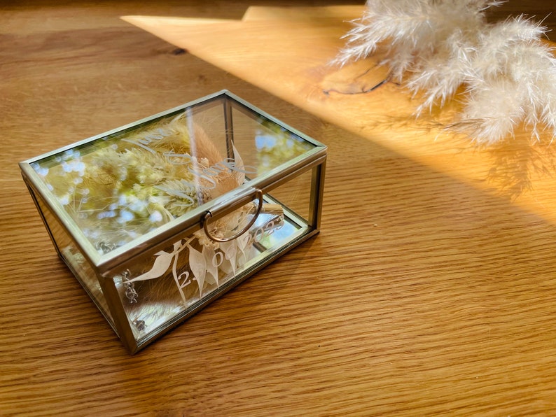 May include: A glass ring box with a gold trim and a floral design. The box is open and the inside is lined with white fabric.