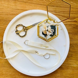 May include: A white round tray with a gold hexagonal ring box with dried flowers inside. The tray also has a pair of gold scissors, a white ribbon with the names "Dunyeya" and "Yase" written on it, and two gold rings.