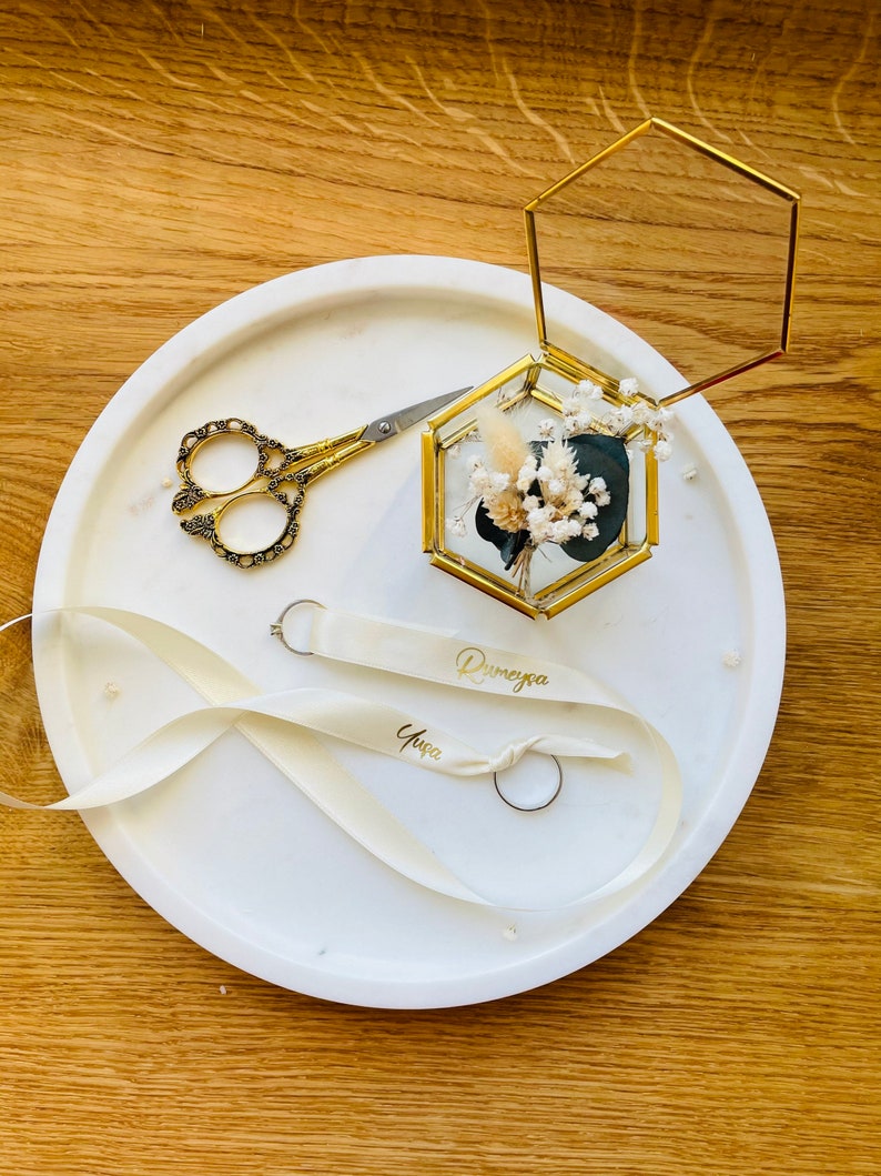 May include: A white round tray with a gold hexagonal ring box with dried flowers inside. The tray also has a pair of gold scissors, a white ribbon with the names "Dunyeya" and "Yase" written on it, and two gold rings.