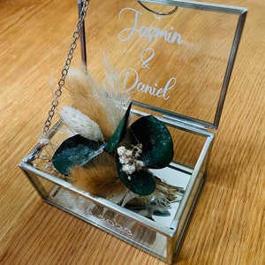 May include: A glass ring box with a silver frame and a hinged lid. The box is decorated with dried flowers and greenery. The lid is engraved with the names "Jasmin & Daniel" and the date "2023".