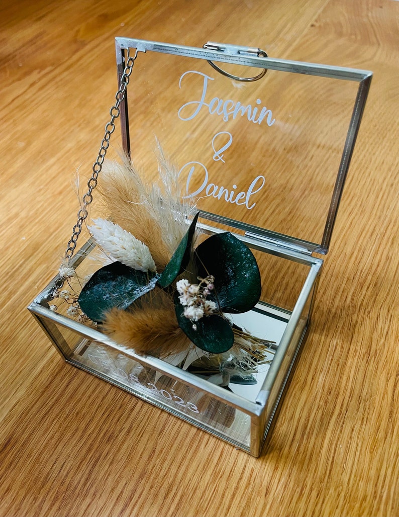 May include: A glass ring box with a silver frame and a hinged lid. The box is decorated with dried flowers and greenery. The lid is engraved with the names "Jasmin & Daniel" and the date "2023".