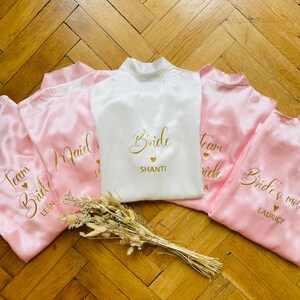 May include: A collection of satin bridal robes in white and pink. The white robe is embroidered with "Bride" and a name in gold. Pink robes are embroidered with "Team Bride" and names. A small bouquet of dried flowers rests in front.