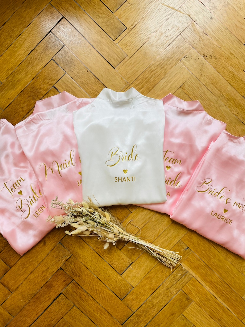 May include: A collection of satin bridal robes in white and pink. The white robe is embroidered with "Bride" and a name in gold. Pink robes are embroidered with "Team Bride" and names. A small bouquet of dried flowers rests in front.