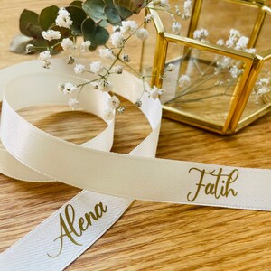 May include: White satin ribbon with the names "Alena" and "Fatih" written in gold lettering. The ribbon is tied around a small, clear glass box with a gold frame.