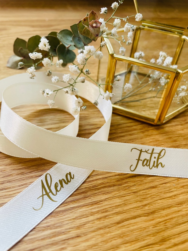May include: White satin ribbon with the names "Alena" and "Fatih" written in gold lettering. The ribbon is tied around a small, clear glass box with a gold frame.